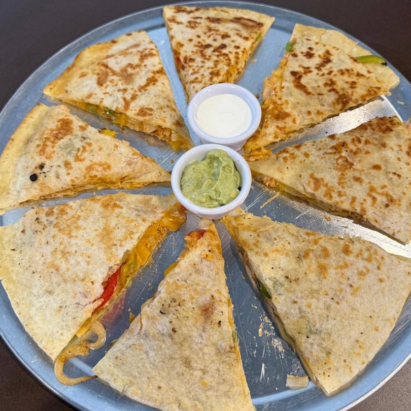 A round metal tray holds eight triangular quesadilla slices with guacamole and sour cream in the center.
