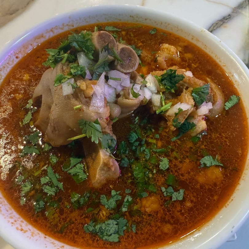 Close-up of a bowl of red menudo soup with large pieces of meat, cilantro, and diced onions.