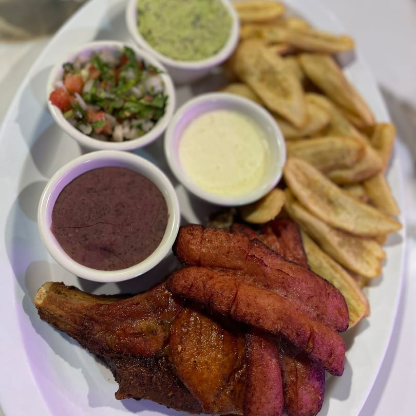 Grilled chicken and sausages with plantain fries, salsa, guacamole, and two sauces on a white plate.