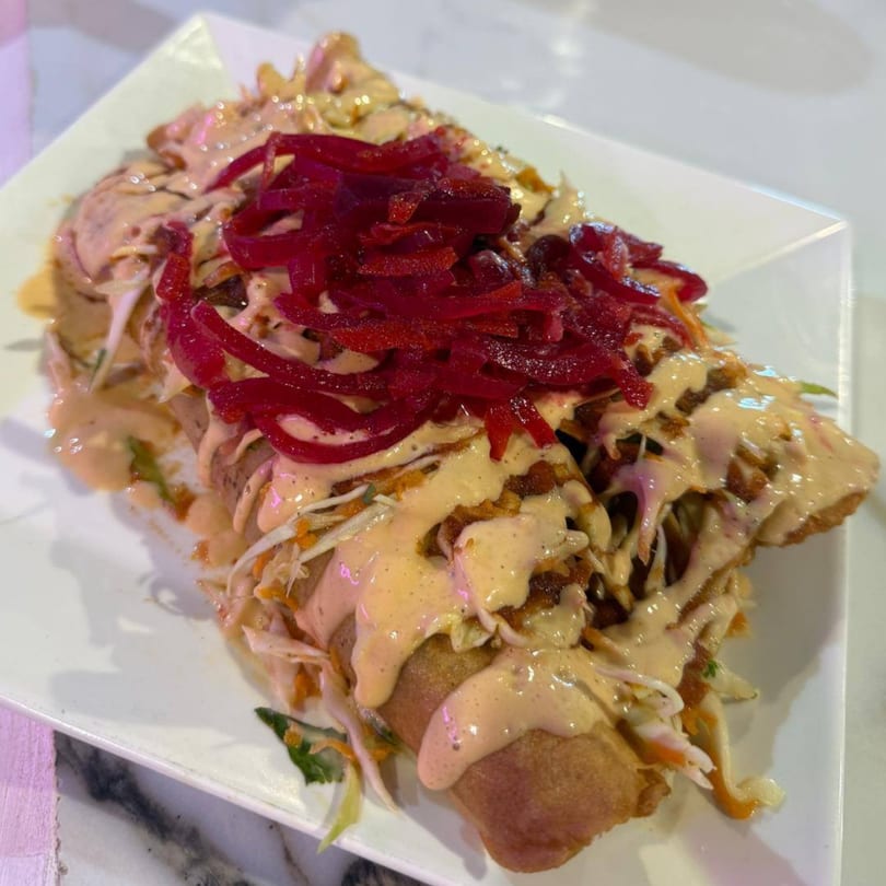 A fried burrito topped with creamy sauce, shredded cabbage, and bright red pickled onions on a white plate.